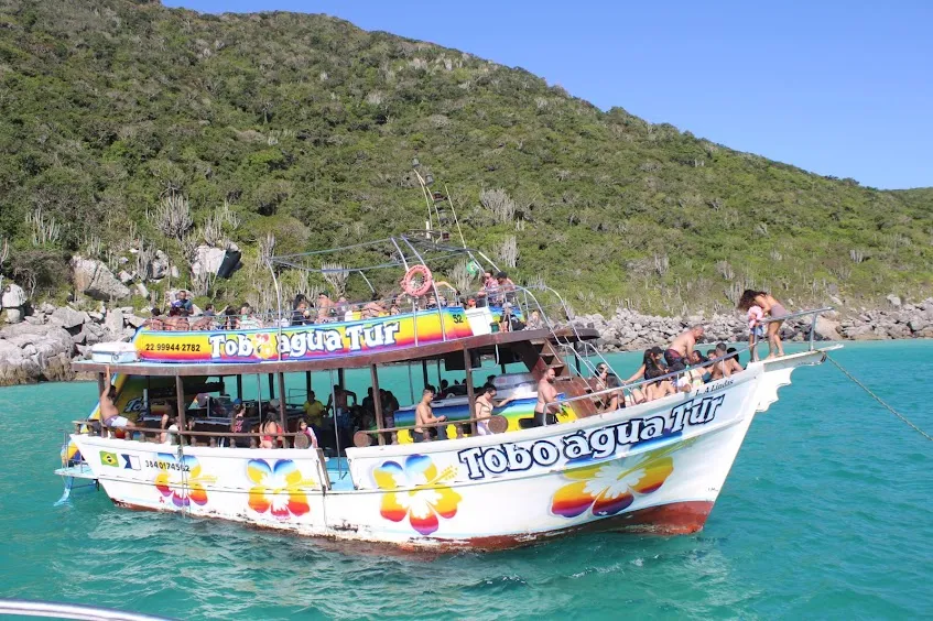Escuna colorida ancorada em água turquesa ao pé de um morro verde, com passageiros no convés e na proa