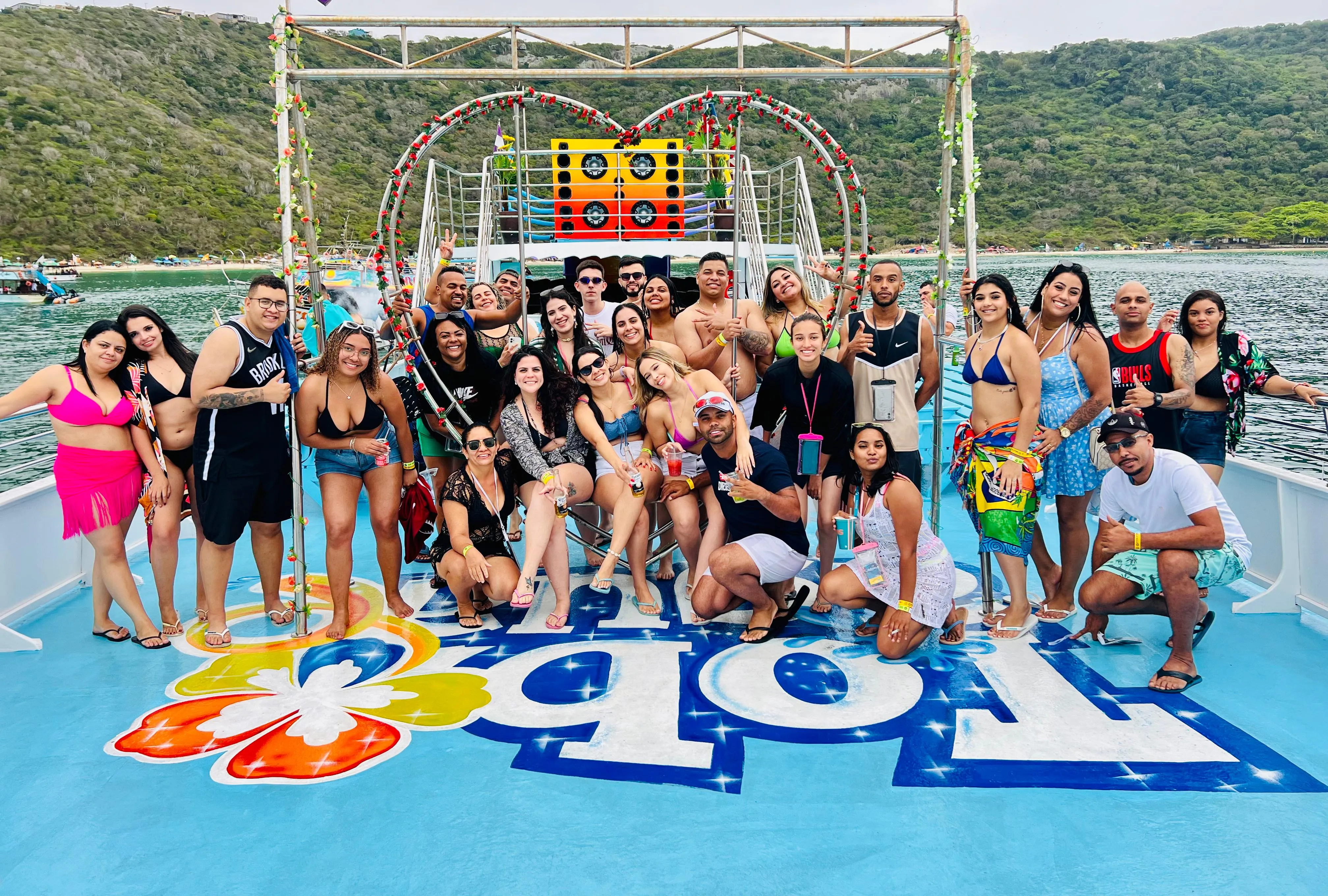 Grupo de passageiros reunidos no convés colorido da escuna, sorrindo para a foto, com o morro verde e o mar ao fundo